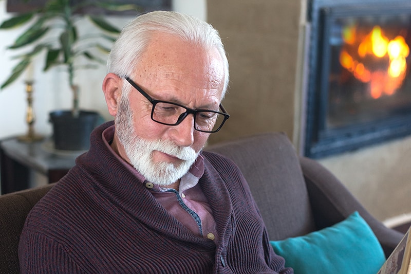 A senior man happily looks at a childhood photo album by the cozy fireplace at The Avenues Crofton Park, a 55+ independent living community that hosts monthly Fireside Chats discussion series