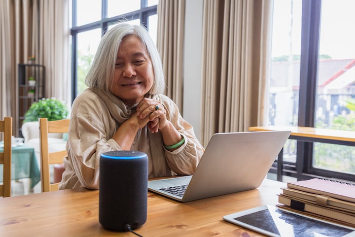 Asian senior woman seated at a dining table with an open laptop computer smiles as she uses smart speaker technology in the comfort of her independent residence at The Avenues Crofton Park