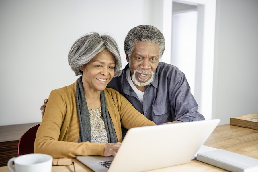 African American senior couple drinking coffee and using a laptop computer to take a virtual retirement community tour from the comfort of home. 